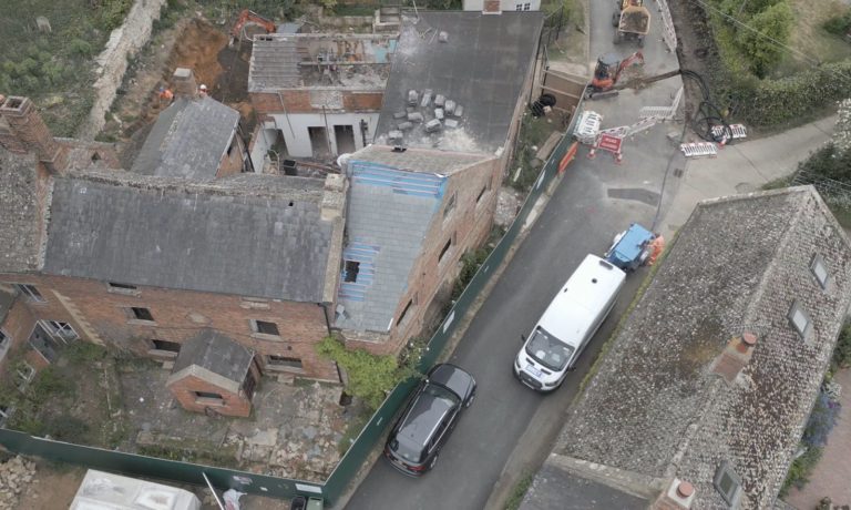 Aerial view of a small road with a white van and black car parked beside old brick buildings; nearby, construction equipment and barriers surround a partially demolished structure.
