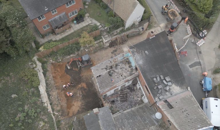 Aerial view of a construction site with workers, a digger, and debris next to houses, roads, and parked vehicles. Some roofs appear damaged or under repair.
