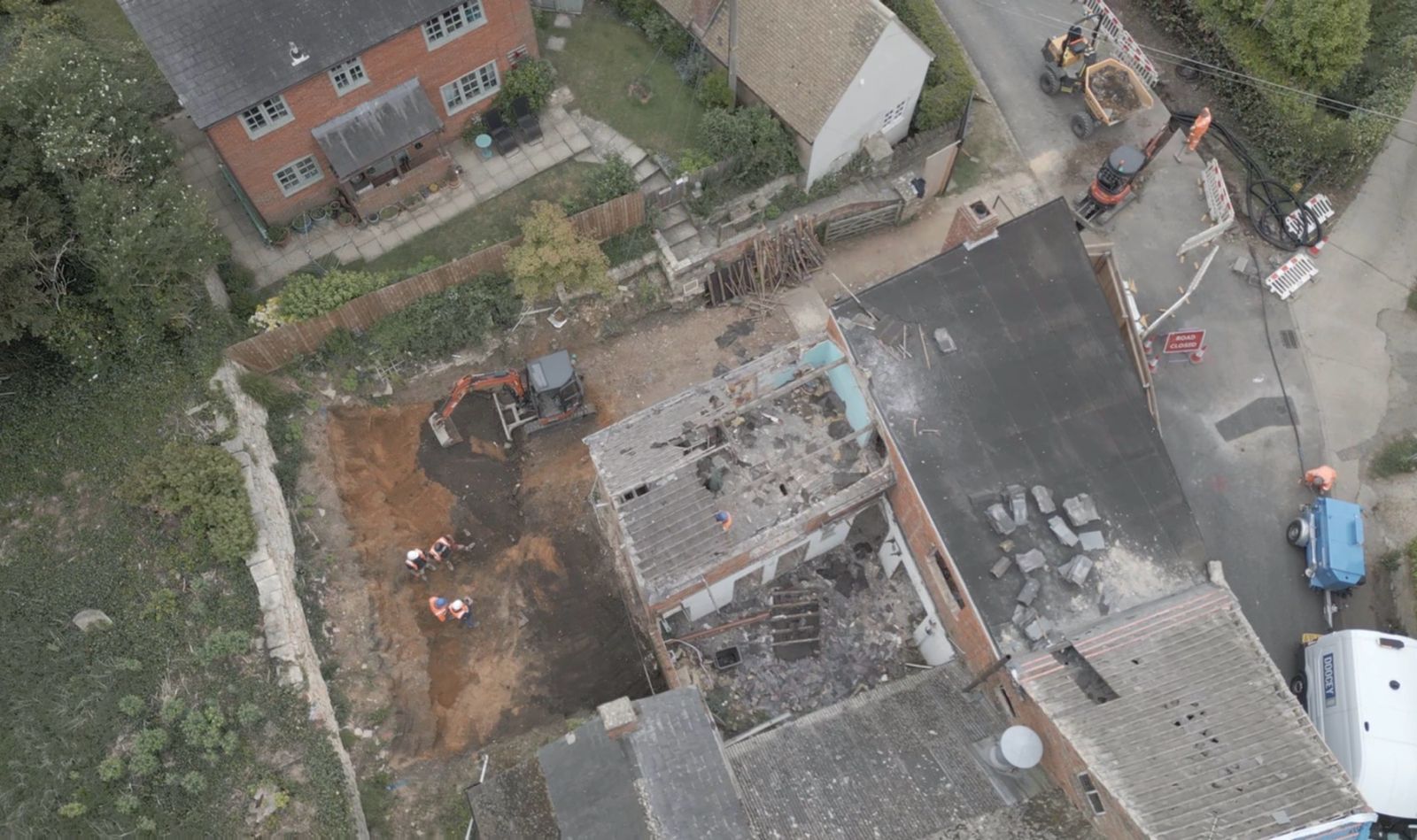 Aerial view of a construction site with workers, a digger, and debris next to houses, roads, and parked vehicles. Some roofs appear damaged or under repair.