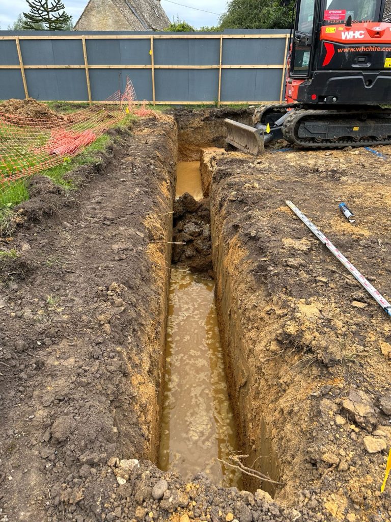 A deep trench filled with muddy water at a construction site, with an excavator nearby and safety fencing along the edge. A measuring tape lies on the ground beside the trench.