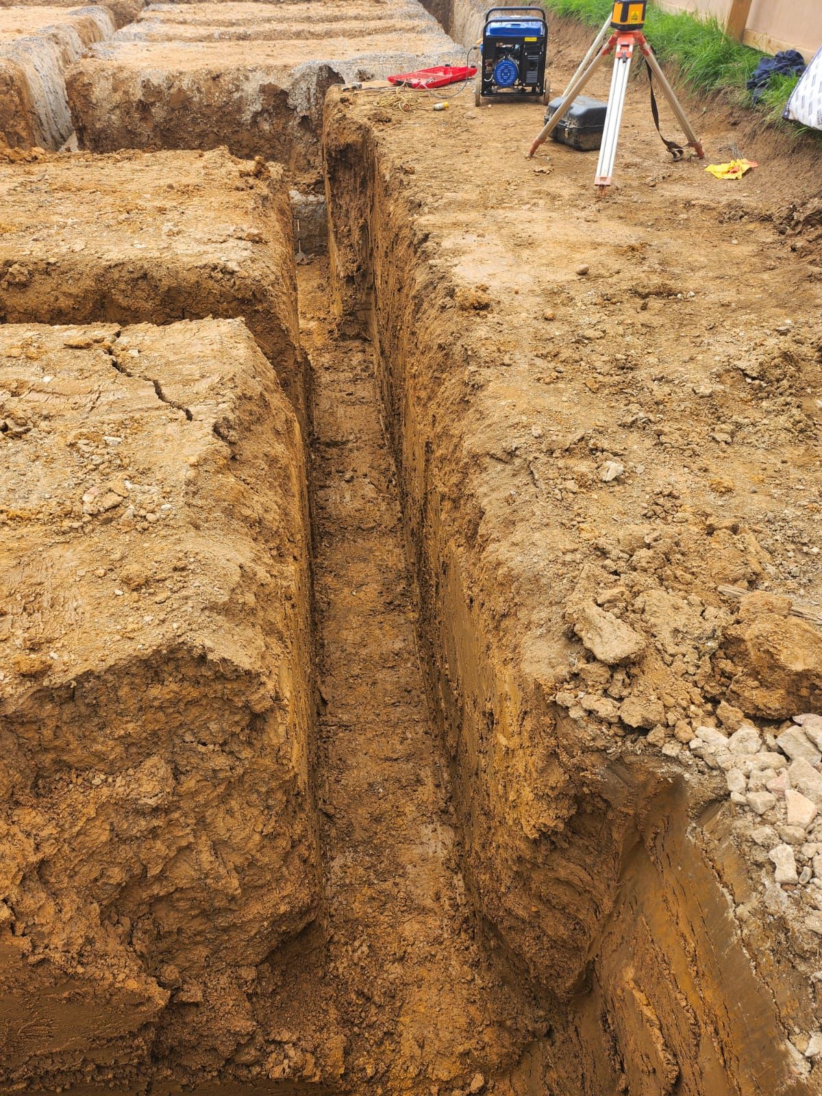 A deep trench dug into brown soil at a construction site, with surveyed dirt walls and equipment like a tripod and generator visible on the surface nearby.