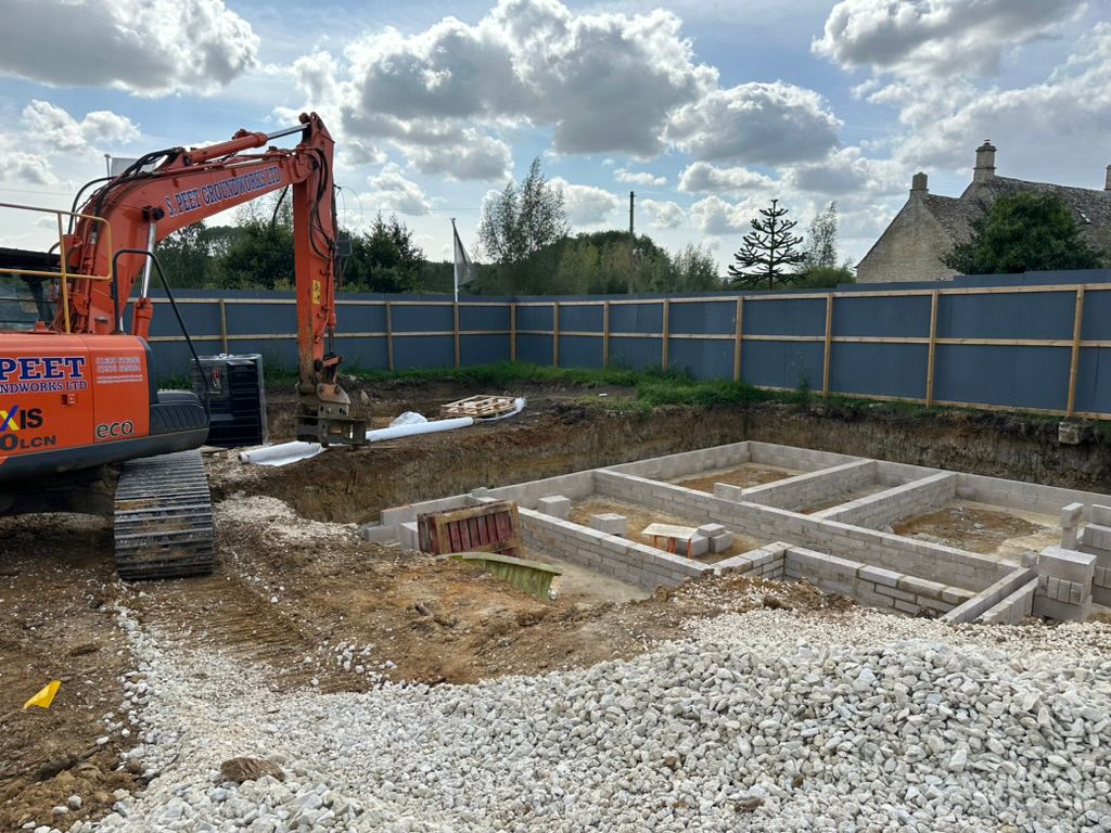 An excavator sits beside a construction site with concrete block foundations in progress, piles of gravel, and wooden fencing surrounding the area. Trees and houses are visible in the background under a partly cloudy sky.