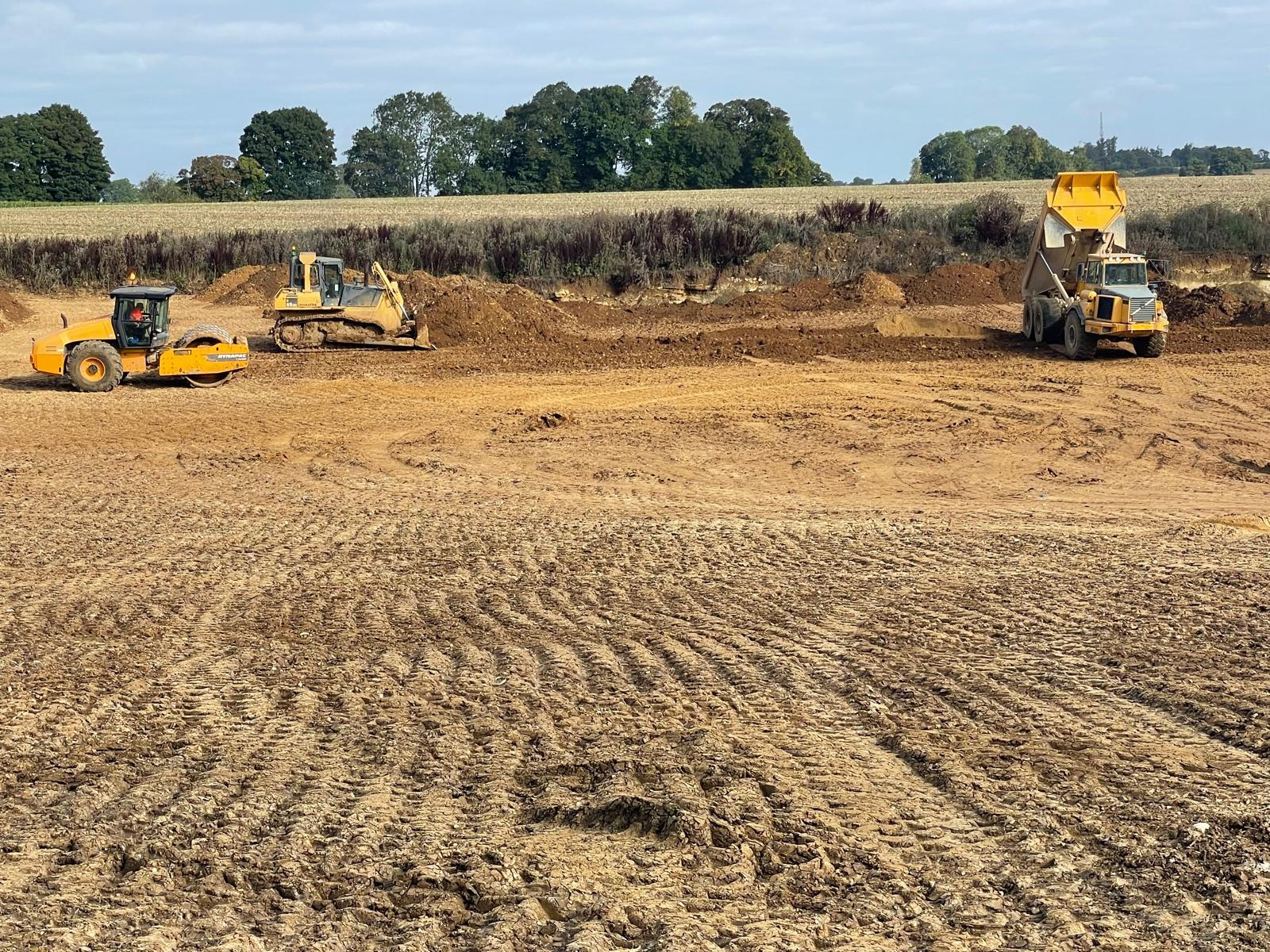 Construction site with a bulldozer, dump truck, and roller working on a large dirt area, surrounded by trees and fields in the background under a partly cloudy sky.