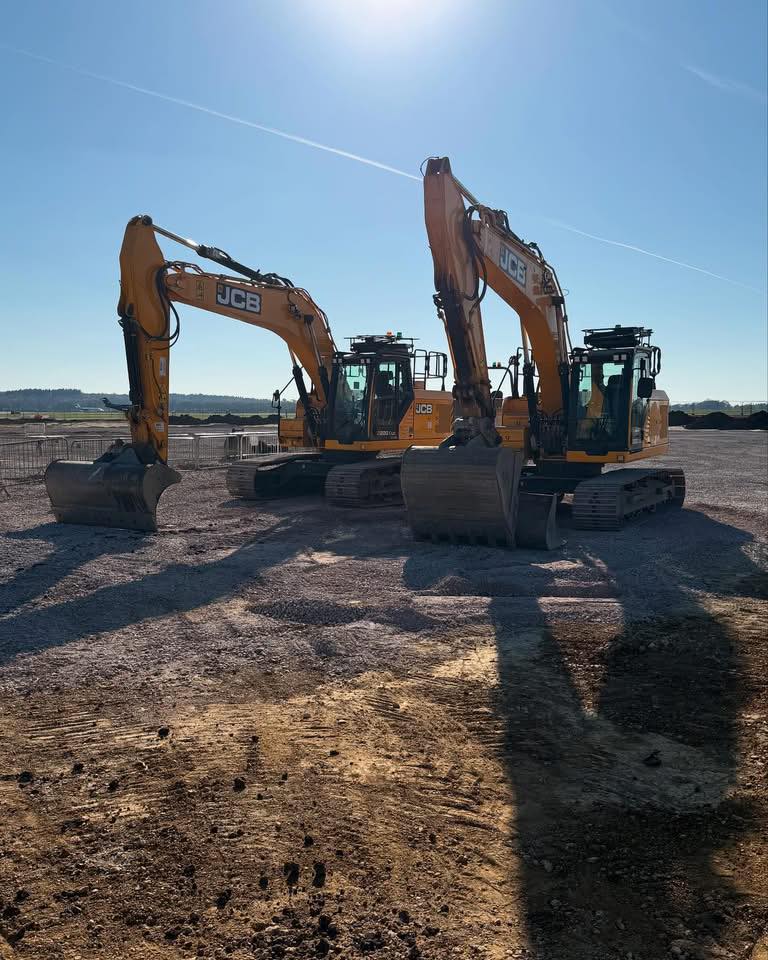 Two yellow JCB excavators are parked on a gravel construction site under a clear blue sky, with sun casting long shadows on the ground.